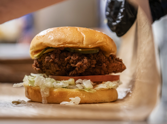 A close-up of a fried chicken sandwich with lettuce, tomato, pickles, and a brioche bun, being wrapped in paper by a person wearing a black glove.